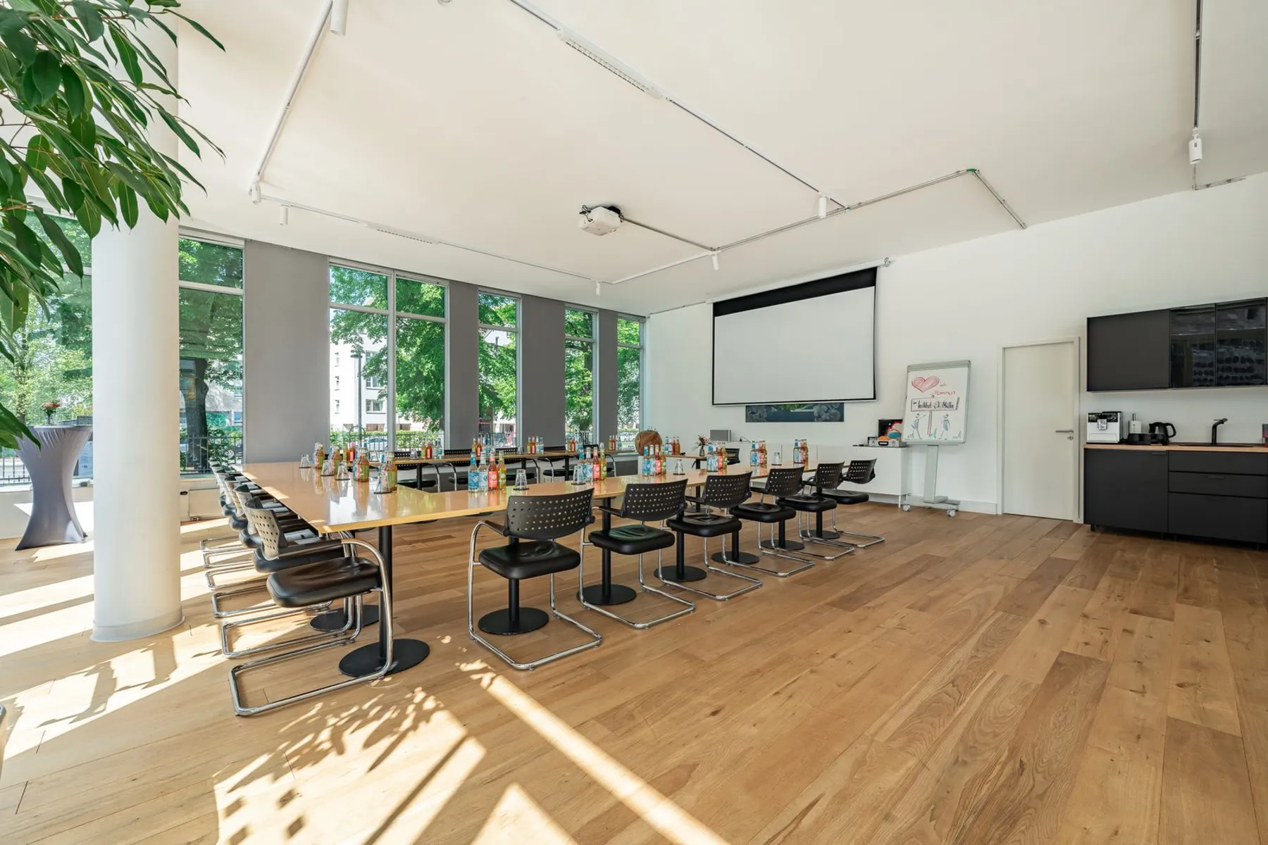 Heller Seminarraum des Instituts mit langer Tischreihe, Freischwingern, Projektionsfläche und bodentiefen Fenstern zu einer baumbestandenen Straße.